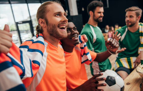 Excited multicultural football fans in orange t-shirts and scarf celebrating victory with ball while their upset friends in different t-shirts sitting behind during watch of soccer match at bar